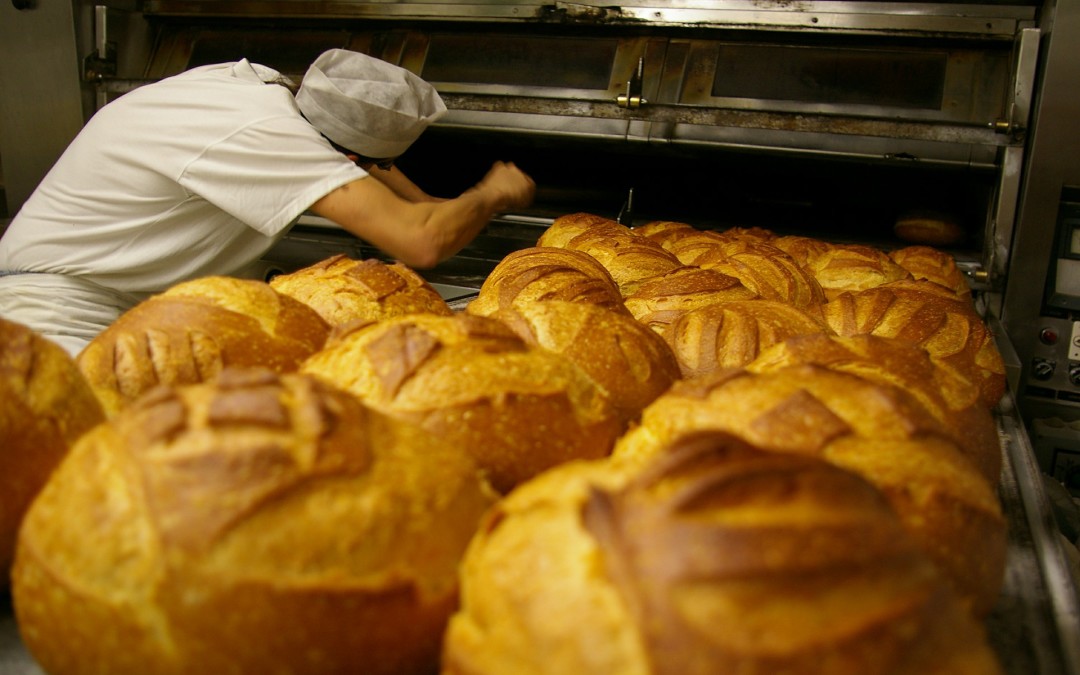 Foreground: Several crusty bread rolls. Background: A baker reaching into an oven.